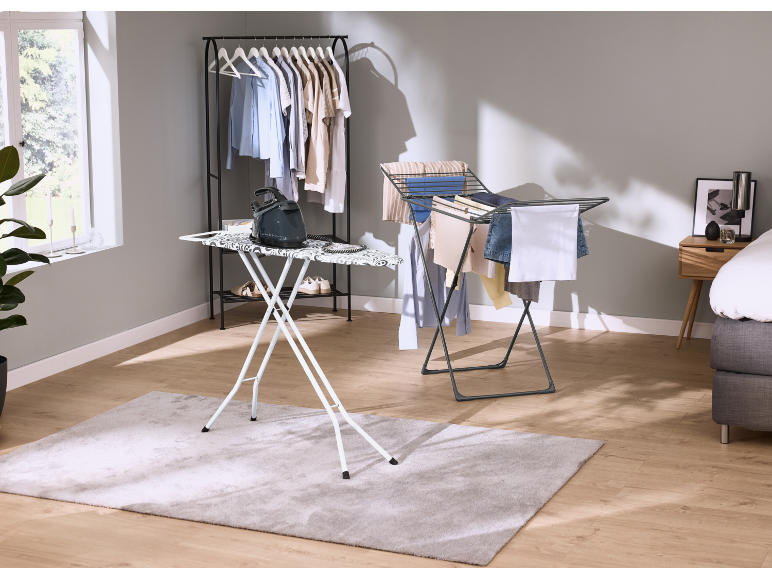 Table à repasser avec fer, portant à vêtements et étendoir dans une pièce.