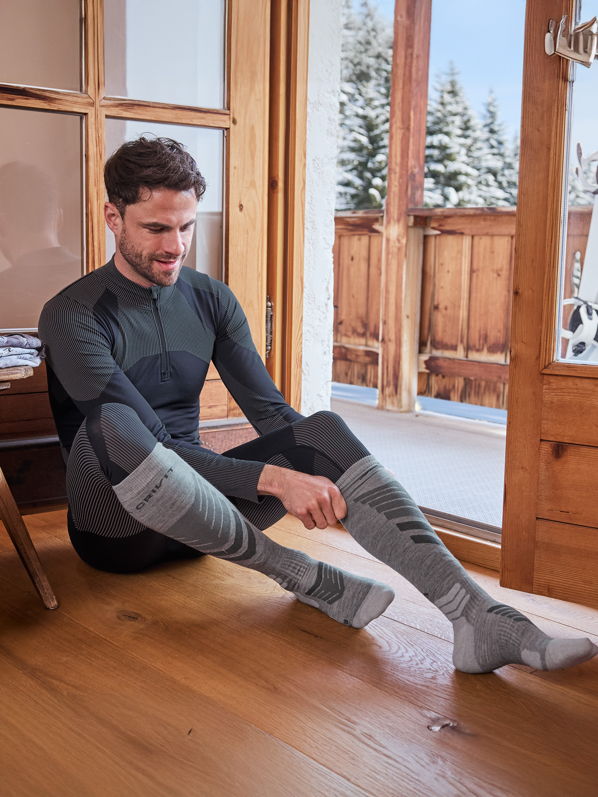 Homme en sous-vêtements thermiques noirs et gris et chaussettes, assis sur un plancher en bois.
