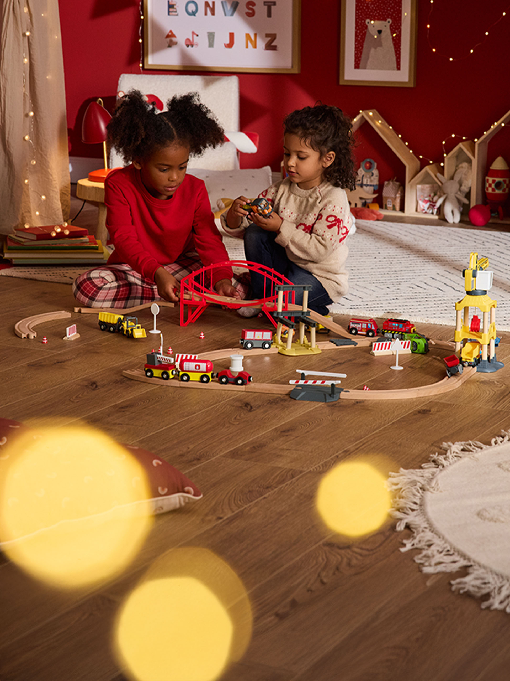 Deux enfants jouent avec un train en bois et des véhicules sur un parquet.