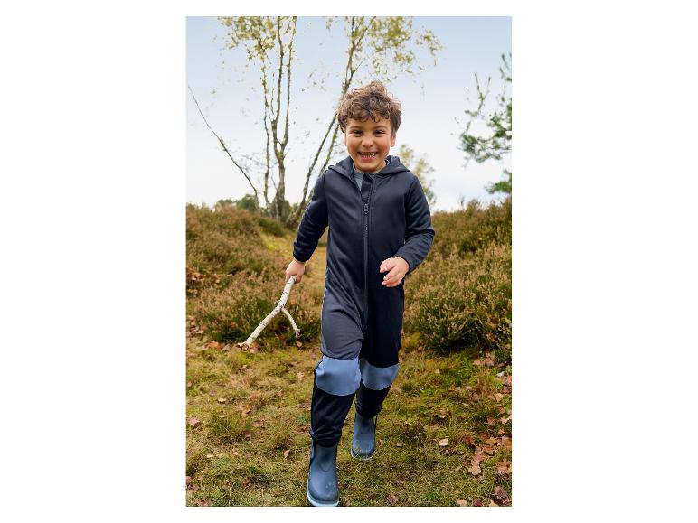 Garçon souriant en combinaison imperméable bleu foncé et bottes, tenant un bâton dans une forêt.