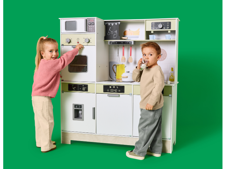 Deux enfants jouant avec une grande cuisine en bois pour enfants sur fond vert.