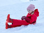 Une fille en combinaison de ski rouge, avec un bonnet rose et des bottes rouges, est allongée dans la neige.