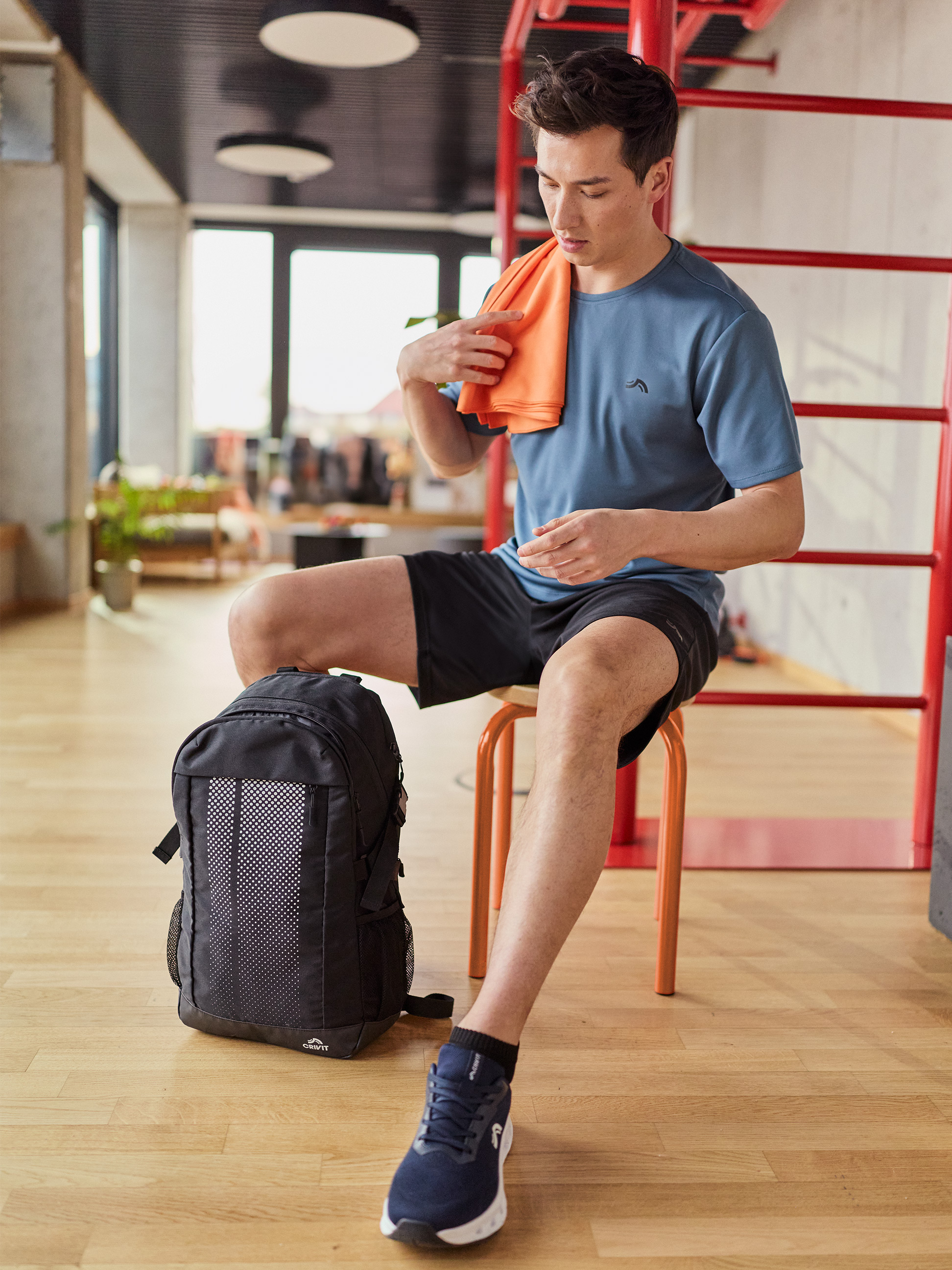 Homme en t-shirt et short de sport, s'essuyant avec une serviette orange, avec un sac à dos à côté.