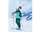 Une femme en tenue de ski verte marche avec des skis sur le dos sur des pentes enneigées.