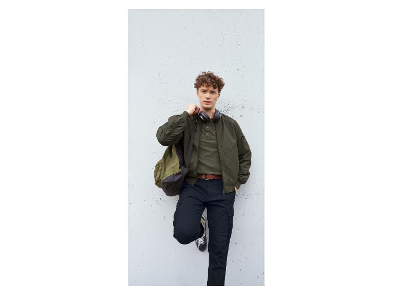 Homme en blouson bomber vert olive, chemise Henley et pantalon cargo foncé avec casque.