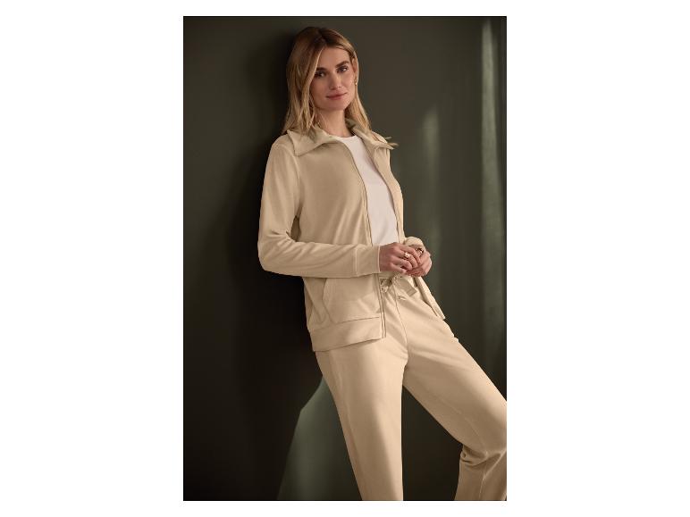 Femme en survêtement beige appuyée contre un mur sombre.