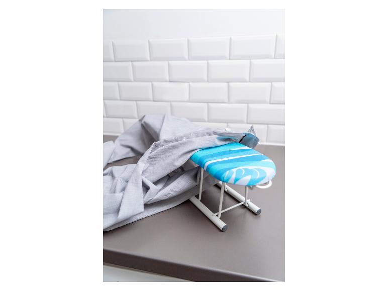 Une mini-table à repasser avec une housse rayée bleu et blanc se trouve sur un comptoir gris avec une chemise grise posée dessus.