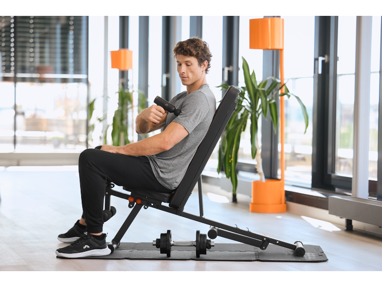 Homme utilisant un pistolet de massage assis sur un banc de musculation dans une salle de sport moderne.