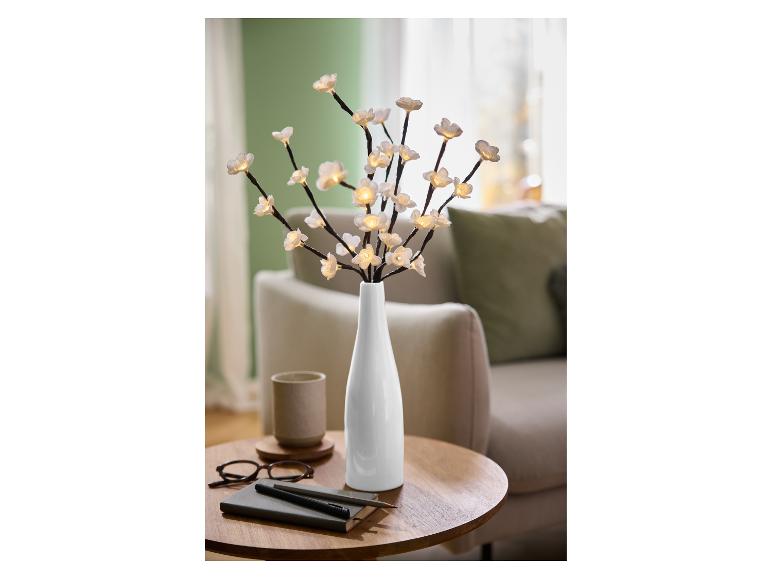 Branches lumineuses LED avec fleurs blanches dans un vase, sur une table en bois avec carnet et lunettes.