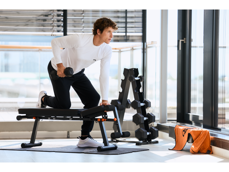 Homme s'entraînant avec un haltère sur un banc de fitness dans une salle de sport avec de grandes fenêtres.