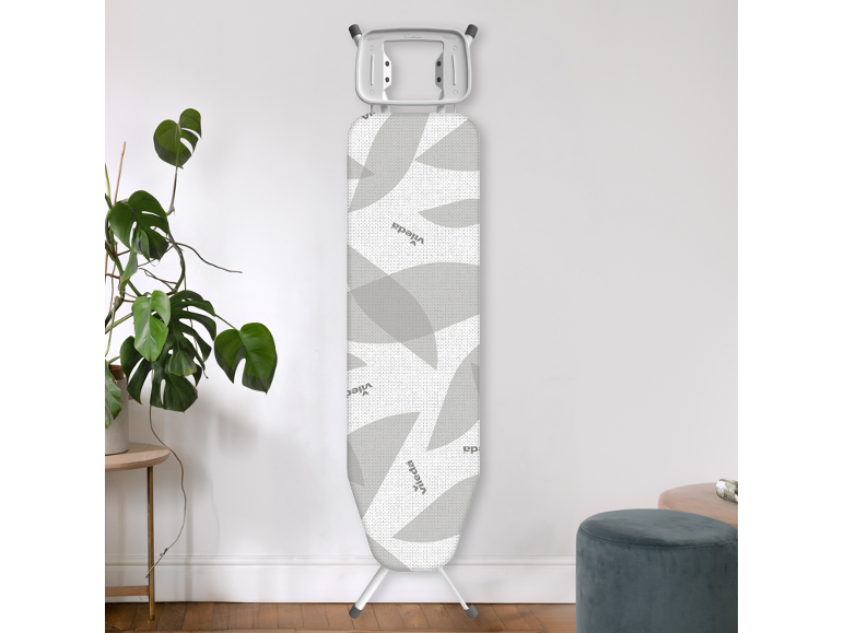 Planche à repasser pliée avec housse à motifs de feuilles grises, posée contre un mur blanc.