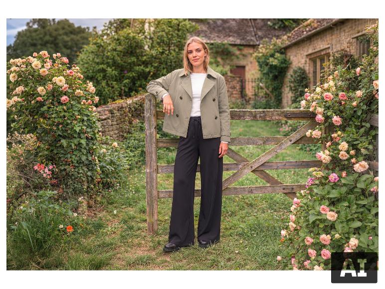 Femme en veste verte courte et pantalon noir large, appuyée sur une clôture en bois.