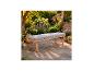 Banc de jardin en bois avec coussin gris sur une terrasse en pierre, entouré de plantes et de fleurs luxuriantes.