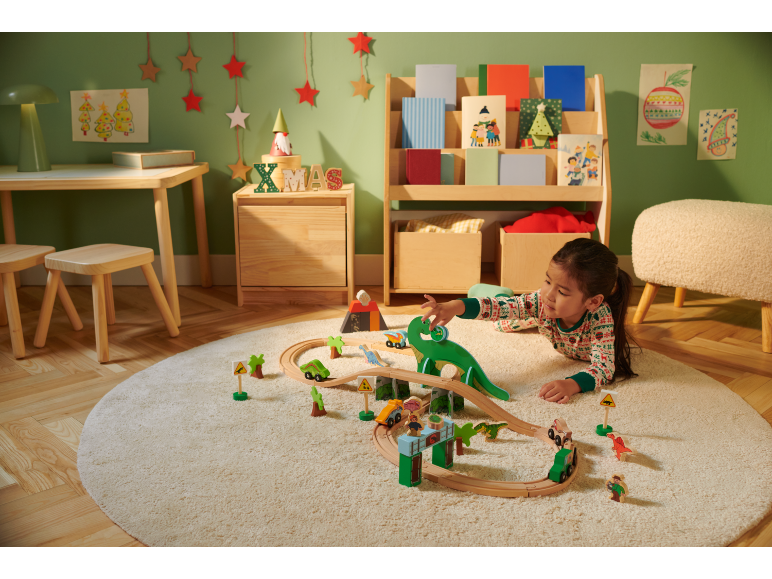 Fille jouant avec un train en bois et des dinosaures sur un tapis rond dans une pièce décorée pour Noël.