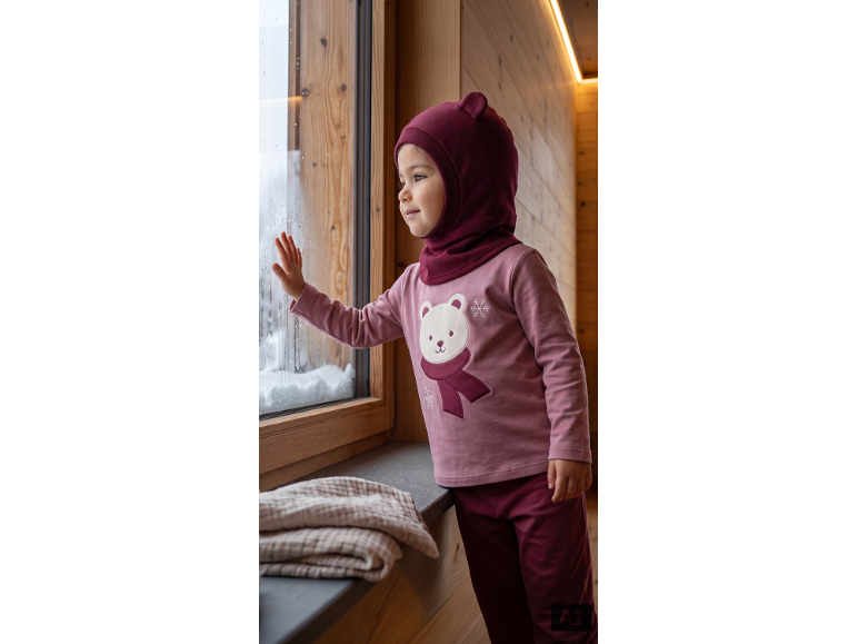 Bébé en pull rose avec ours et cagoule bordeaux regarde la neige par la fenêtre.