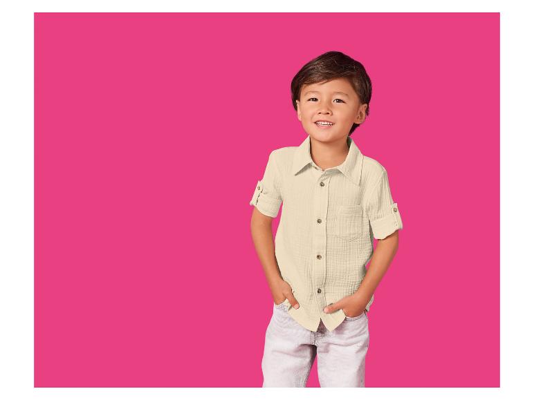 Garçon souriant en chemise crème et pantalon clair sur fond fuchsia.