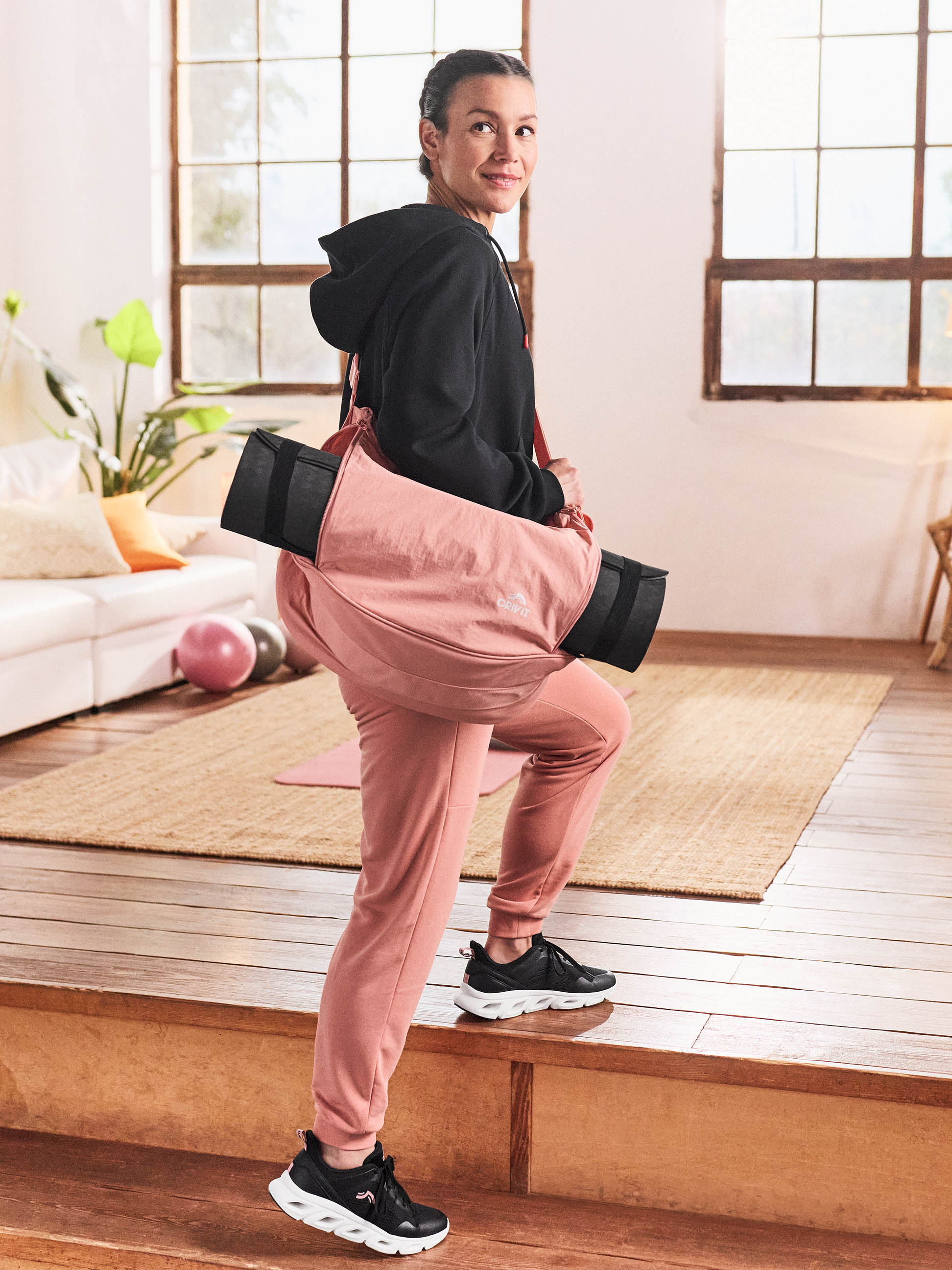 Femme en sweat à capuche noir et pantalon de jogging rose avec un tapis de yoga sur l'épaule.