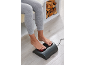 Femme utilisant un masseur de pieds électrique chauffant, assise sur un banc avec du bois de chauffage.