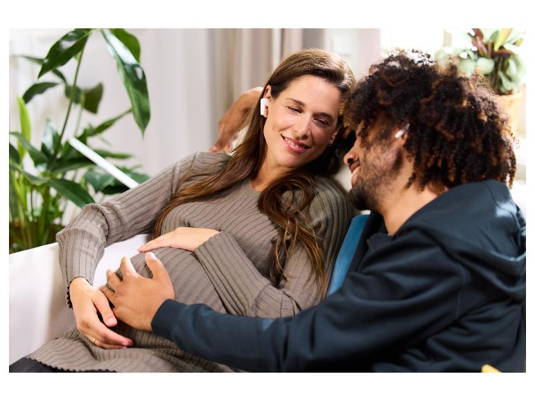Une femme enceinte et son partenaire avec des écouteurs sans fil sont assis sur le canapé.