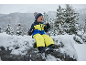 Enfant en combinaison de ski jaune et bleue, bonnet et gants, assis sur un rocher enneigé.