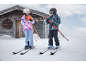 Deux enfants en tenue de ski avec skis et bâtons dans un paysage enneigé près d'une cabane en bois.