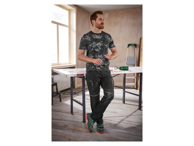 Un homme porte des vêtements de travail, dont un t-shirt camouflage, un pantalon noir et des chaussures de sécurité vertes.