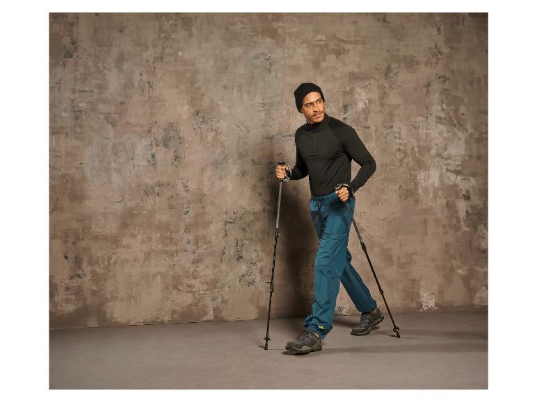Homme avec des bâtons de randonnée et des chaussures de randonnée marchant devant un mur en béton.