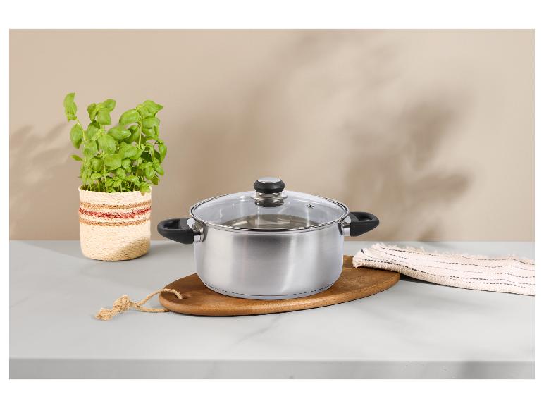 Casserole en acier inoxydable avec couvercle en verre, herbes fraîches et planche en bois sur comptoir en marbre.