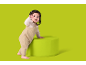 Bébé en salopette beige debout à côté d'un pouf vert, regardant l'appareil photo.