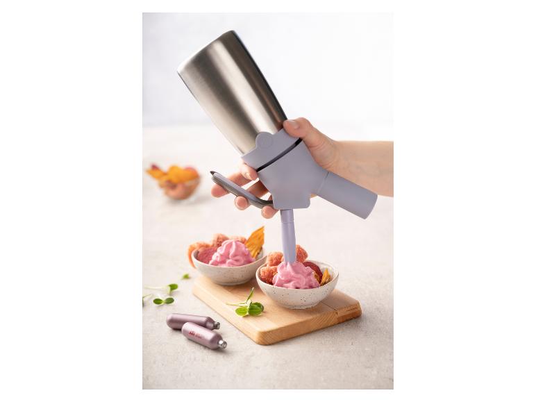 Une main utilise un siphon de cuisine pour verser de la mousse rose dans des bols de fruits secs.