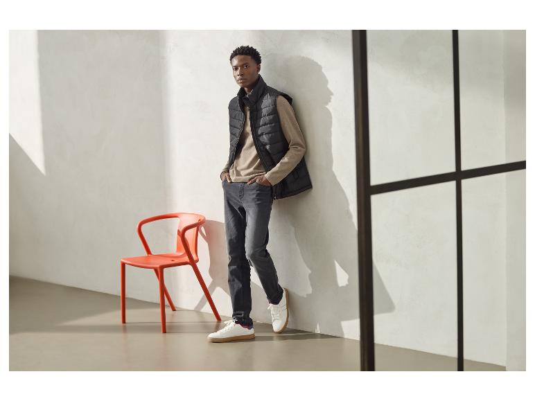 Homme en gilet matelassé noir, chemise beige à manches longues et jean gris, appuyé contre un mur.