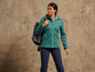 Une femme en veste softshell verte et pantalon de randonnée bleu avec un sac à dos.