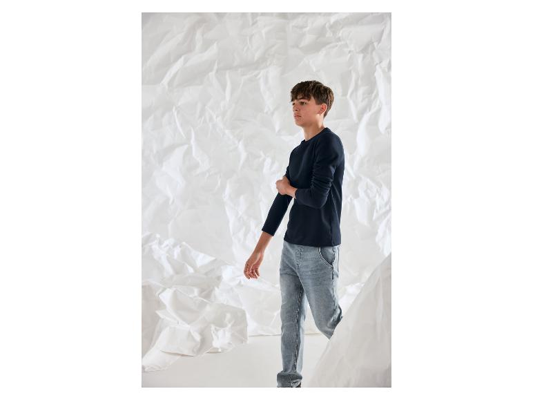 Un garçon en T-shirt bleu foncé à manches longues et jean clair devant un fond blanc froissé.