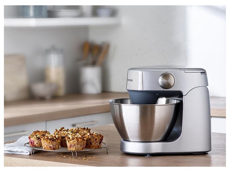 Robot de cuisine argenté avec bol en acier inoxydable et muffins sur une table en bois.
