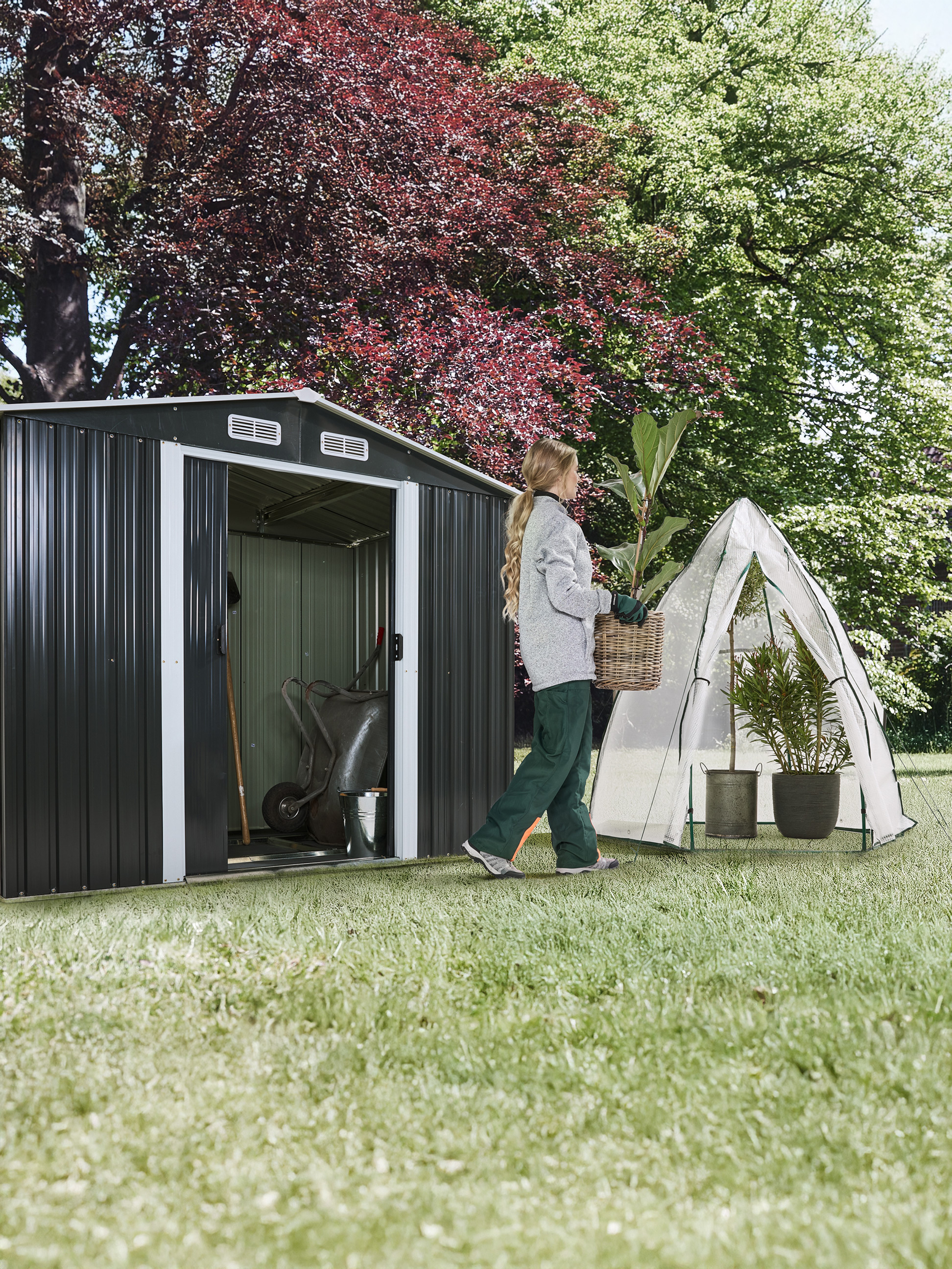 Femme transportant une plante dans un panier, à côté d'un abri de jardin et d'une petite serre.