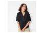 Femme en chemise noire à manches courtes et pantalon beige, souriante et regardant à droite.