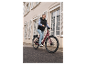 Femme souriante conduisant un vélo électrique rouge bordeaux dans la rue.