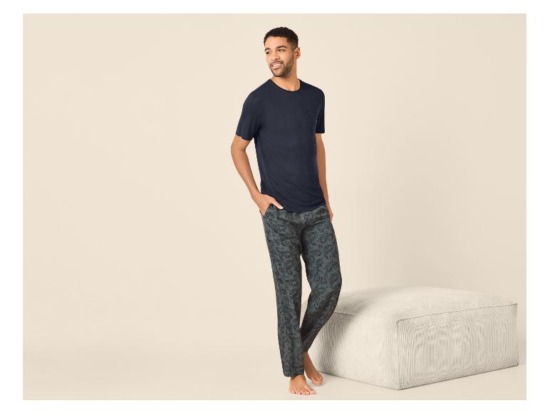 Homme en T-shirt bleu foncé et pantalon de pyjama foncé à motifs, debout près d'un pouf.