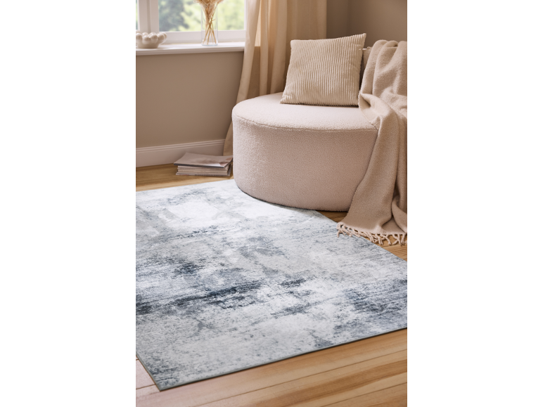 Tapis gris et blanc sur parquet avec pouf et coussin beiges.