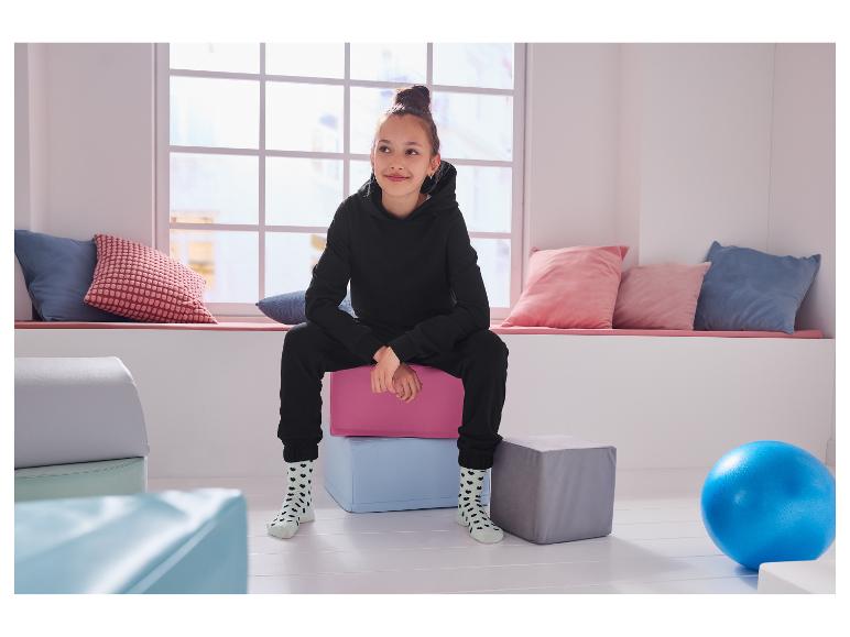 Une fille souriante assise sur des cubes moelleux, portant un sweat à capuche noir et des chaussettes à cœurs.