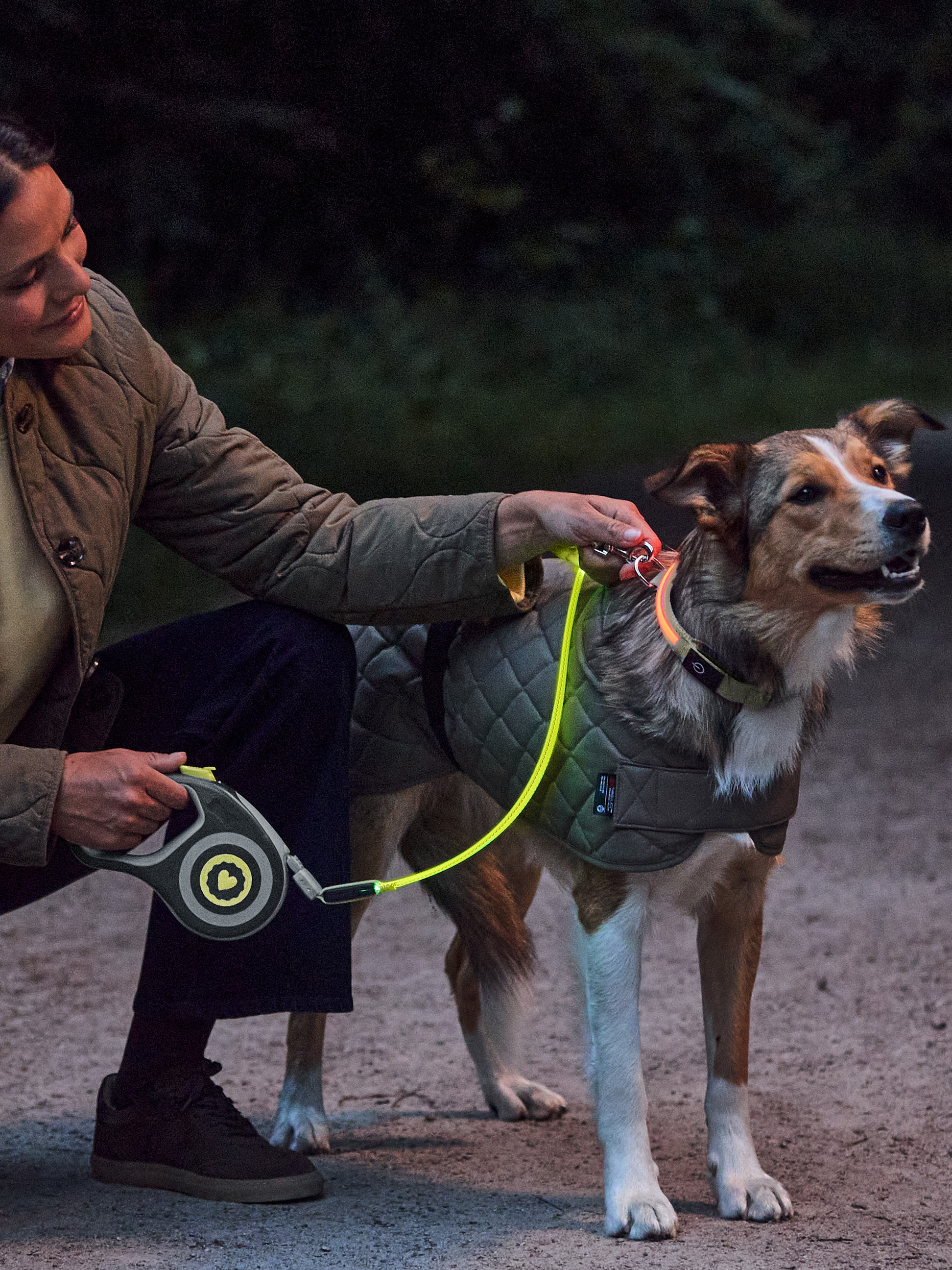 Femme avec chien portant un manteau, un collier et une laisse réfléchissants la nuit.