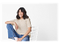 Une femme souriante porte un t-shirt beige et un jean, assise sur une chaise blanche.