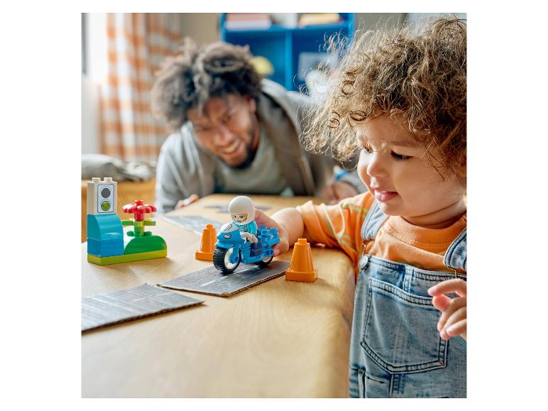 Enfant joue avec une moto jouet et des panneaux de signalisation sur une table, père en arrière-plan.