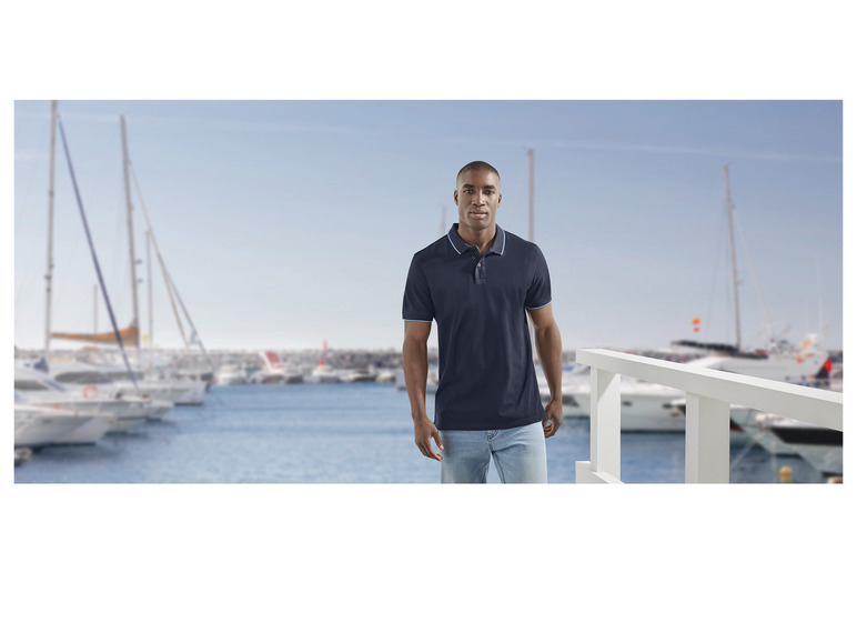 Homme en polo bleu marine et en jean, debout sur un quai avec des bateaux en arrière-plan.