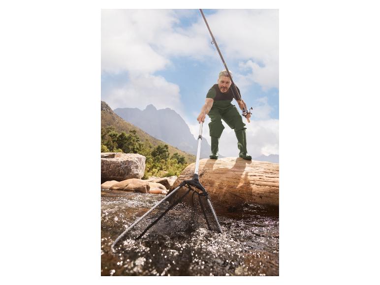 Pêcheur en combinaison imperméable avec canne et épuisette dans un ruisseau de montagne.