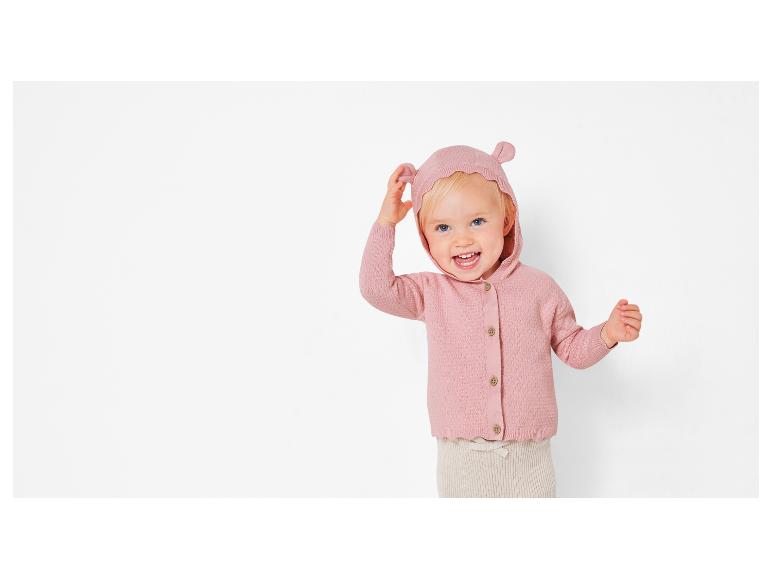 Un bébé portant un pull rose en tricot à capuche et un pantalon beige.