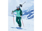 Un skieur en tenue de ski verte, avec des skis sur l'épaule et des bâtons à la main.