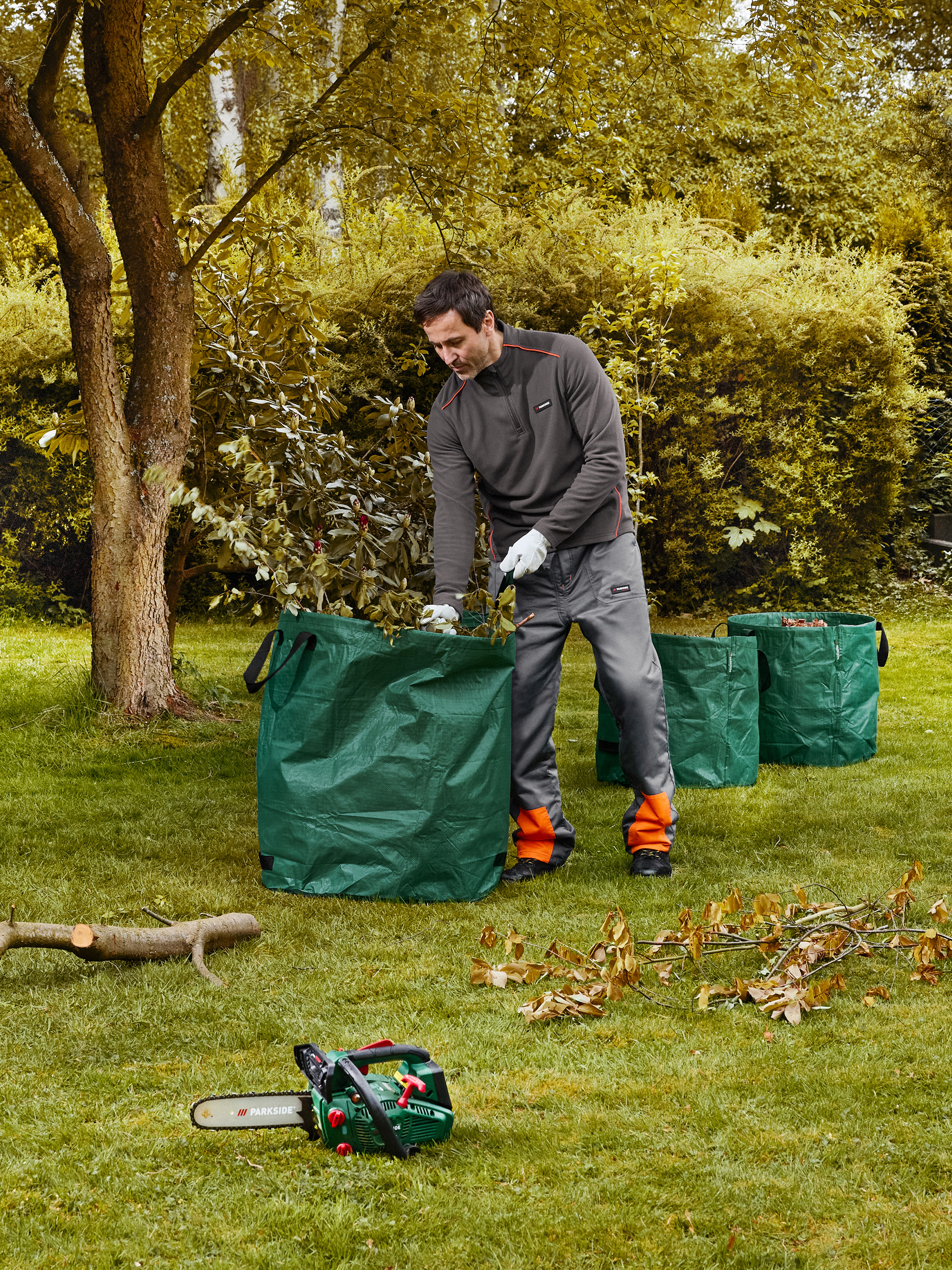 Homme ramassant des branches dans des sacs de jardin, avec une tronçonneuse au sol.