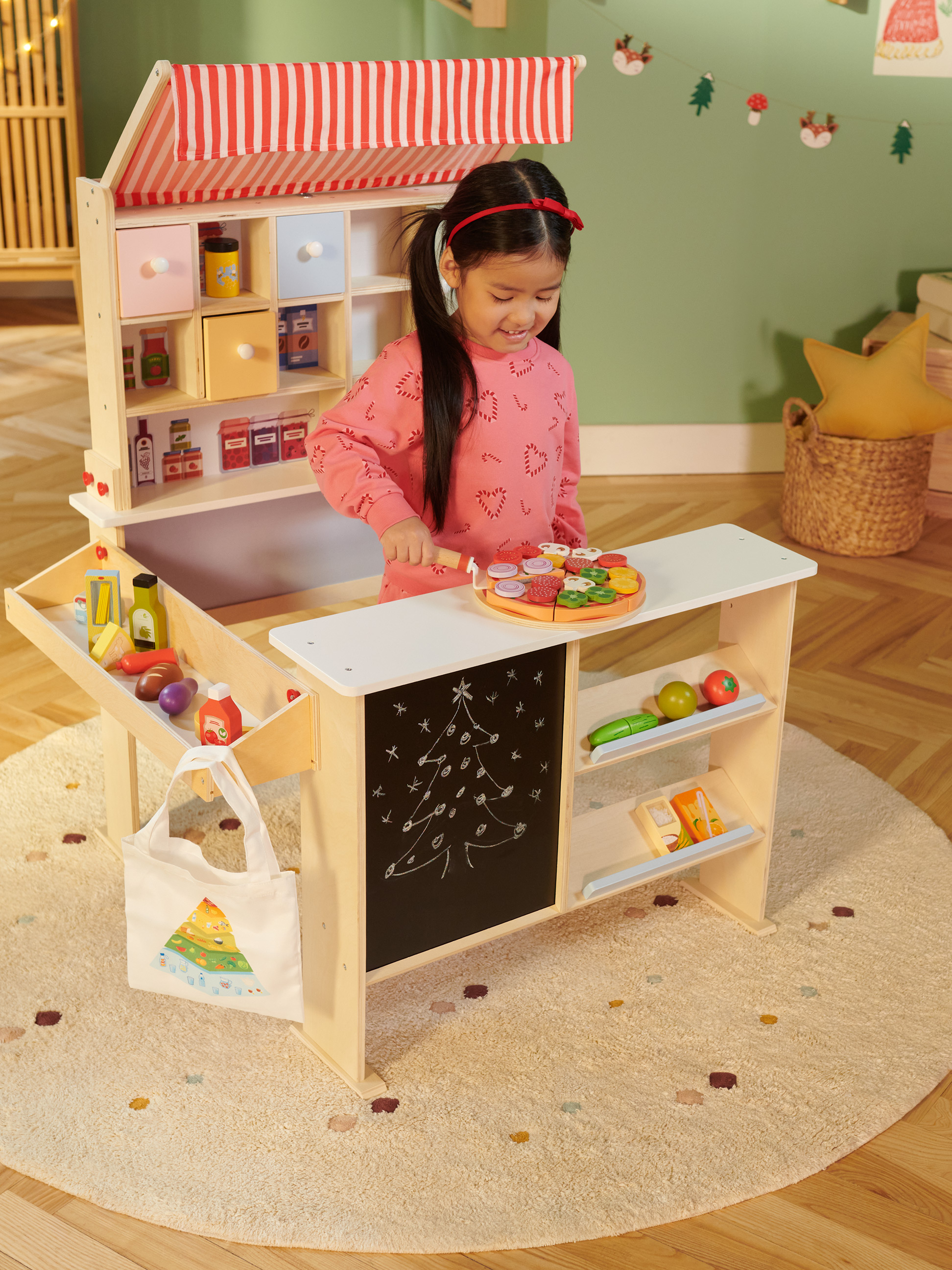 Fille souriante joue avec un stand de magasin de jouets en bois, équipé de nourriture jouet et d'un tableau noir.