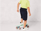 Un enfant portant un t-shirt vert et un short bleu est debout sur une planche à roulettes.